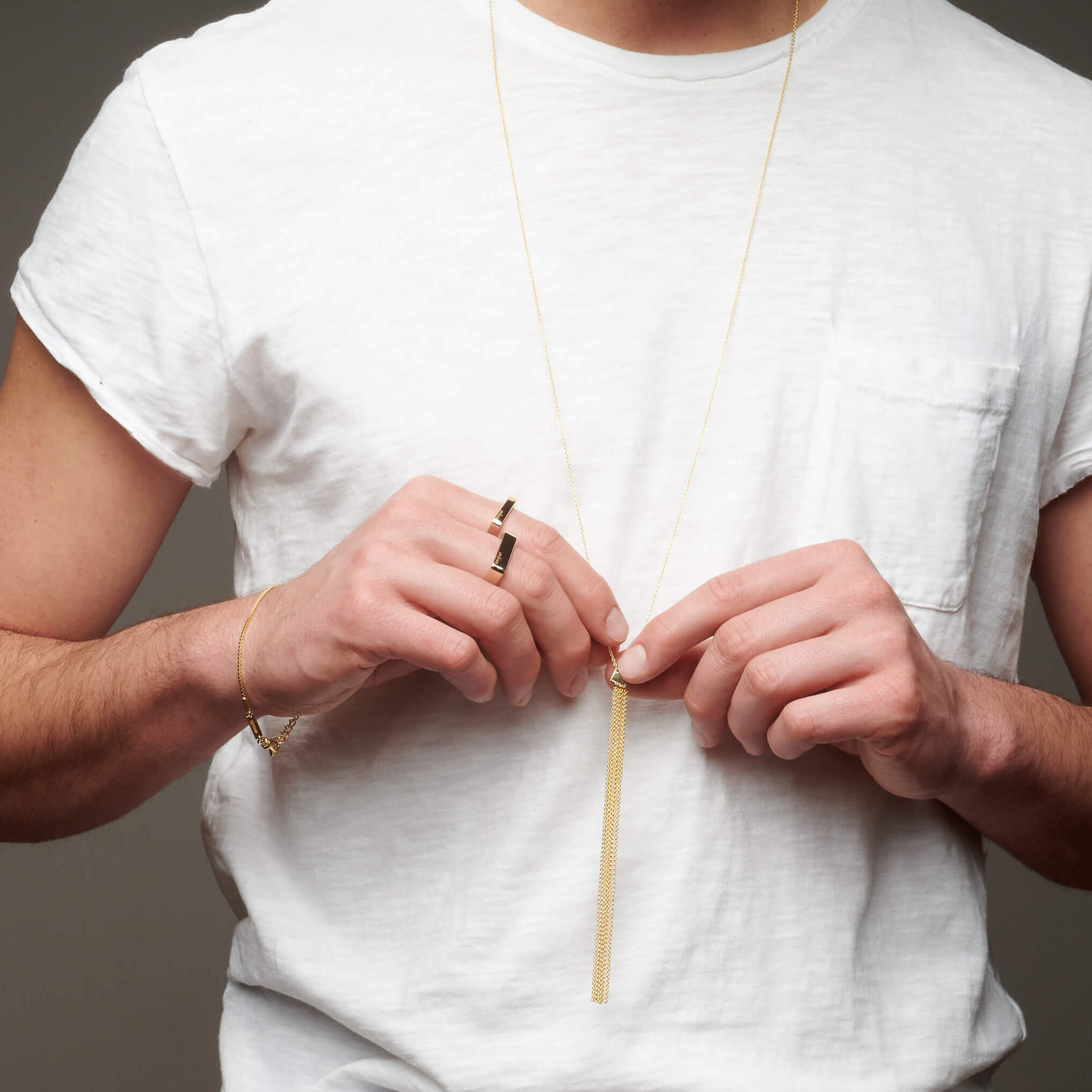 Man's torso with hands holding long atelium necklace wearing gold bracelet and rings