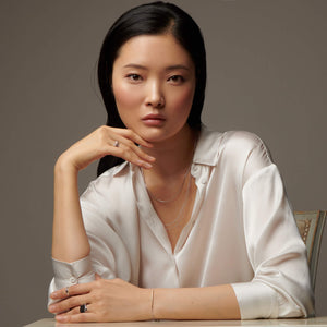 Woman sitting facing forward with her hands by her face wearing atelium bracelet, rings, and necklace