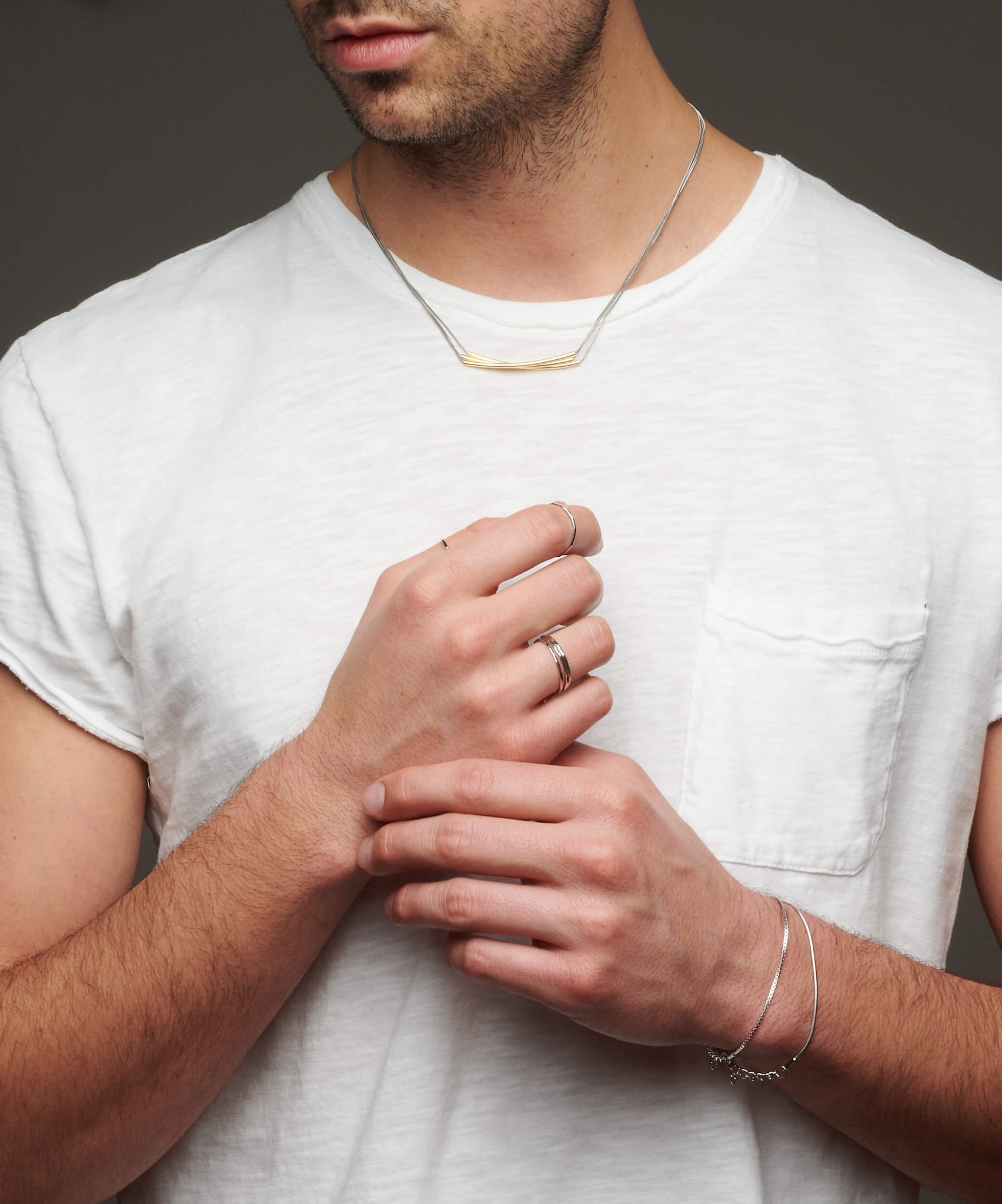 Man's torso with hands together wearing multiple pieces of jewelry including silver atelium bracelet, silver rings, and necklace